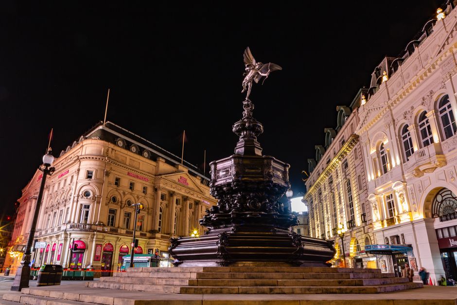 Nighttime view of Shaftesbury Avenue, showing a historic square with ornate buildings illuminated by exterior lighting. At the centre of the scene is a large, dark metal monument with a multi-tiered b