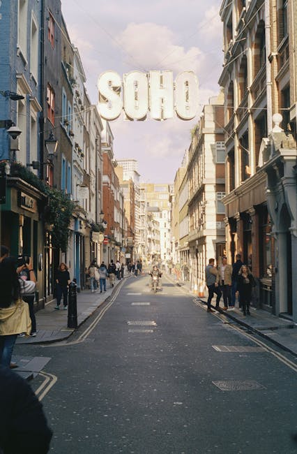 Nighttime view of Shaftesbury Avenue, showing a historic square with ornate buildings illuminated by exterior lighting. At the centre of the scene is a large, dark metal monument with a multi-tiered b